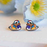 A pair of hand-painted wooden earrings depicting a multicolored mandarin duck design, placed on a white surface with a flower in the background.